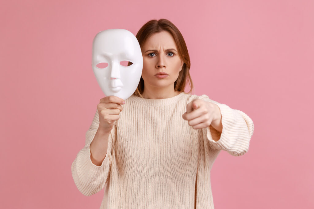 Portrait of strict bossy blond woman pointing finger to camera and holding white mask in her hand, wants to change personality, wearing white sweater. Indoor studio shot isolated on pink background.