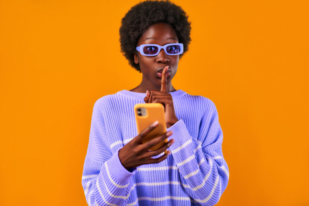 African American young woman with afro hair styling standing in a blue sweater and glasses on a bright orange background using the phone and showing a gesture of quietly placing a finger to her mouth.